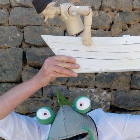 Ein Mann mit Froschmaske hält über seinem Kopf ein kleines, weißes Holzboot, in dem eine Marionette aus Holz sitzt und ihn fragend anschaut.